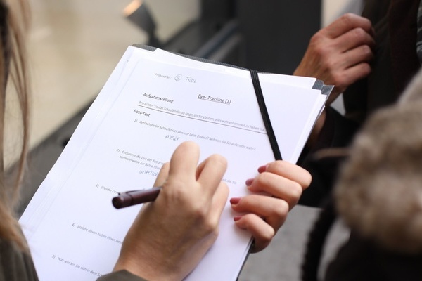 Eine Person mit langen Haaren mit Stift in der Hand fllt ein Formular auf einem Klemmbrett aus- gegenber steht eine Person, die von ihr befragt wird