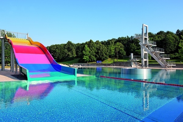 Ansicht Freibad mit farbiger Rutsche und Sprungturm im Hintergtund, dahinter groe Bume