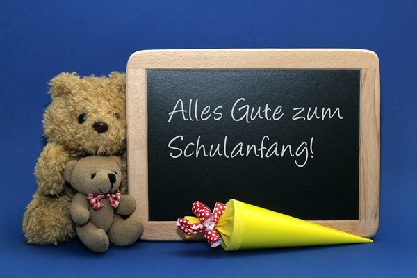 Teddybären und eine schwarze Tafel mit der weißen Aufschrift: Alles Gute zum Schulanfang!. Davor liegt eine gelbe Schultüte.