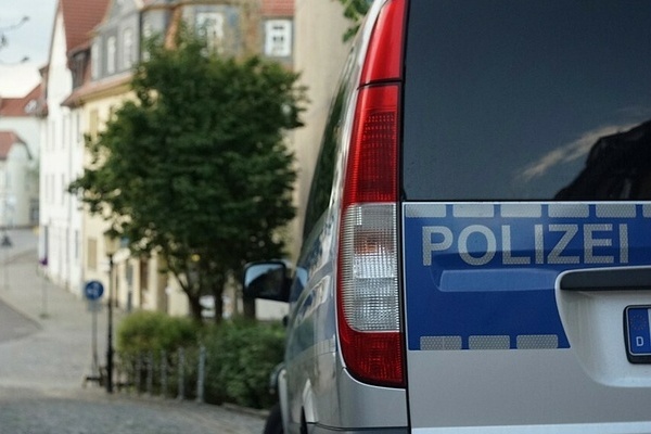 Auf dem Bildausschnitt ist die Rückseite eines Polizeiautos und der Schriftzug Polizei in Grau auf blauem Untergrund. In der Ferne sieht man verschwommen eine Straße und einen Baum sowie eine Häuserreihe.
